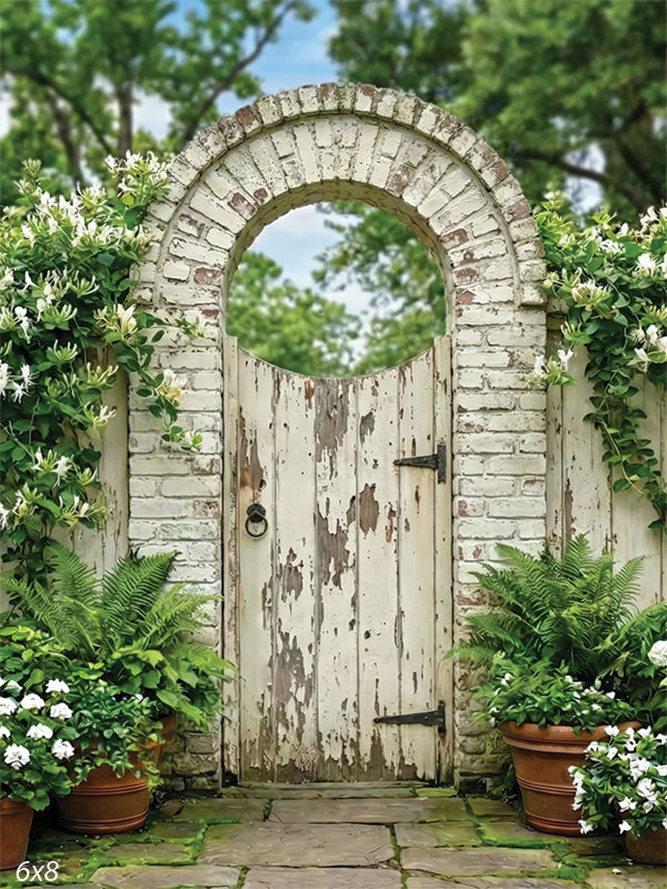 A studio background showing a white brick garden arch with a weathered wooden gate, climbing vines, potted plants, and a stone walkway used as a photography backdrop.