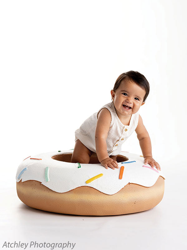 Baby leaning forward in a playful pose on a large sprinkled donut photography prop, shot against a white studio backdrop with soft lighting.