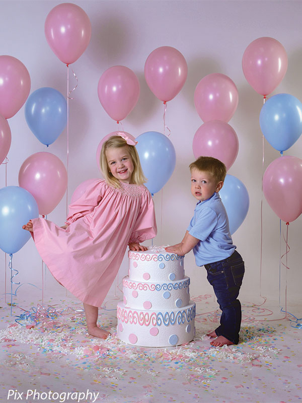 Young girl and boy pose together with blue and pink balloons and a double-sided cake prop.