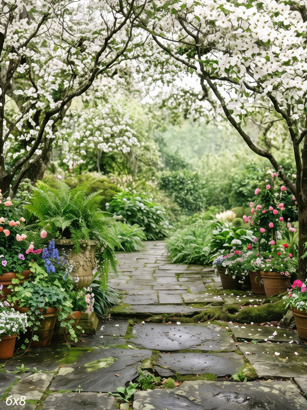 Stone garden path beneath blooming white dogwood trees with ferns, hostas and potted pink roses, a floral scenic photography backdrop.