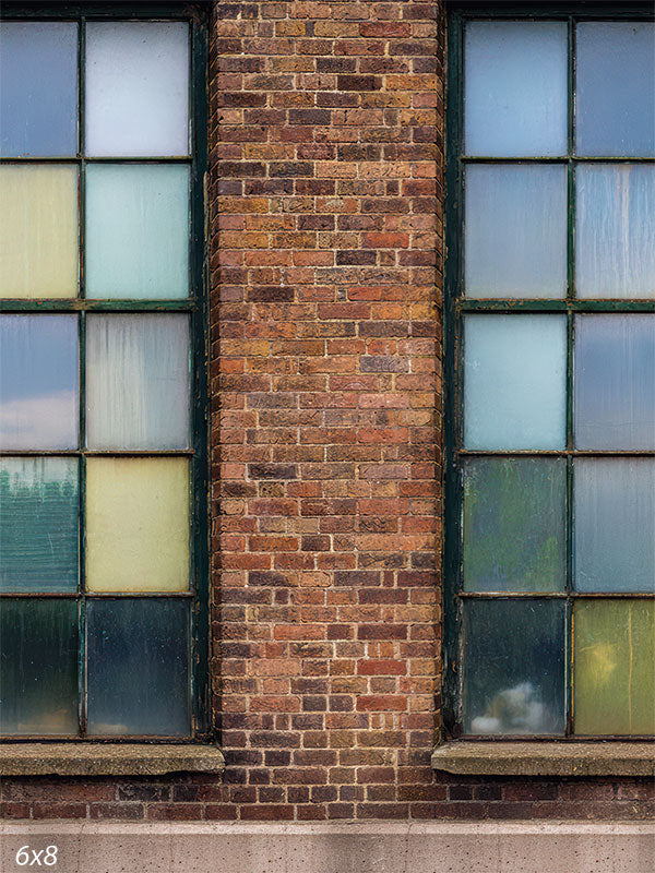 Printed photography backdrop showing a brick warehouse wall with large multi-pane industrial windows and mixed frosted, tinted glass sections. The window grids and brick texture create an urban architectural background behind a subject.