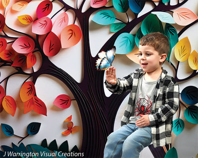 Child with butterfly on hand sits near red, teal, and orange paper leaf tree with white background.
