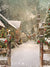 Charming Snow-Covered Holiday Village Backdrop - Charming snow-covered holiday village street with quaint wooden cottages adorned with garlands, wreaths, and twinkling lights, framed by rustic wooden posts and featuring a tall church steeple in the background under a snowy sky.