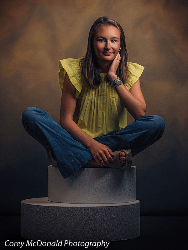 Circular foam posing pedestals used as studio seating, with a senior girl in a yellow ruffle-sleeve top and blue flared jeans seated cross-legged on a single pedestal with a hand at her cheek against a warm brown backdrop.