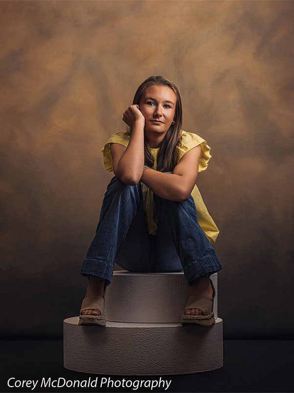Circular foam posing pedestals used as studio seating, with a senior girl in a yellow ruffle-sleeve top and blue flared jeans seated on the pedestal with her knees drawn up and chin on her hand against a warm brown backdrop.