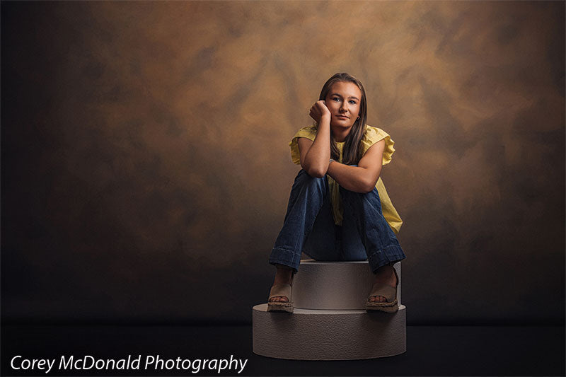 Circular foam posing pedestals used as studio seating, with a senior girl in a yellow ruffle-sleeve top and blue flared jeans seated on the pedestal with her knees drawn up in a wide studio view against a warm brown backdrop.