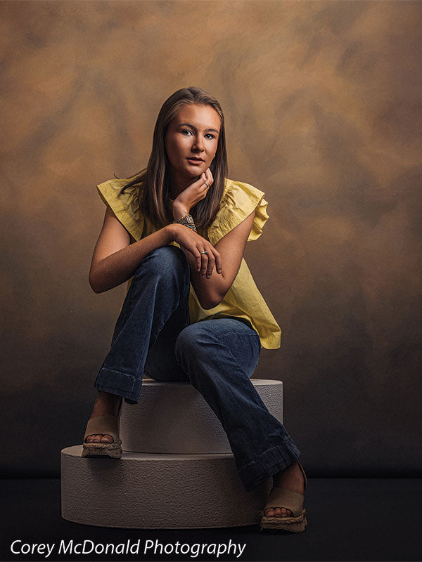Circular foam posing pedestals used as studio seating, with a senior girl in a yellow ruffle-sleeve top and blue flared jeans seated on the stacked pedestals with a hand at her cheek against a warm brown backdrop.