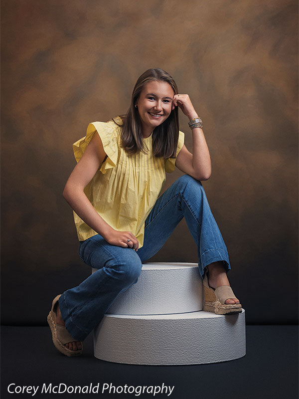 Circular foam posing pedestals used as studio seating, with a senior girl in a yellow ruffle-sleeve top and blue flared jeans seated on the stacked pedestals with a hand resting on her knee against a warm brown backdrop.