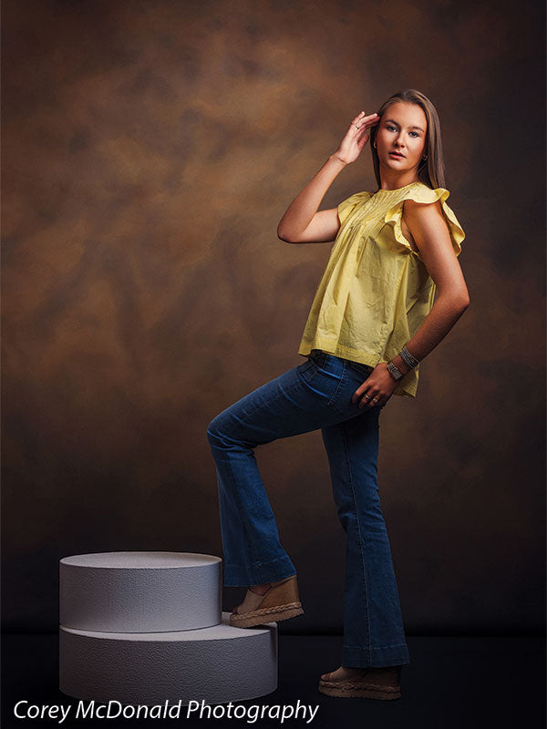 Circular foam posing pedestals used as studio seating, with a senior girl in a yellow ruffle-sleeve top and blue flared jeans standing with one foot on the pedestal and a hand in her hair against a warm brown backdrop.