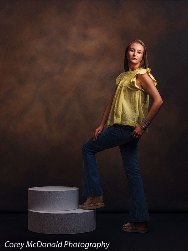 Circular foam posing pedestals used as studio seating, with a senior girl in a yellow ruffle-sleeve top and blue flared jeans standing with one foot resting on the low stacked pedestals against a warm brown backdrop.