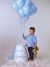 Toddler boy with teddy bear poses beside cake prop and blue balloons on seamless light gray studio backdrop.