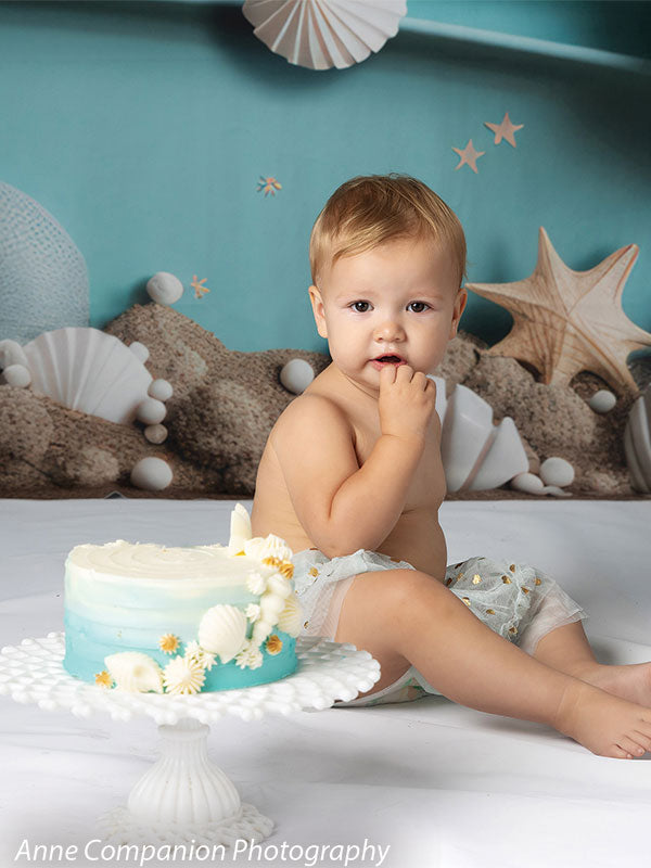 Elegant coastal-themed cake smash backdrop featuring seashells, starfish, a sandy base, and a serene aqua blue background with hanging paper lanterns for a dreamy beach celebration.