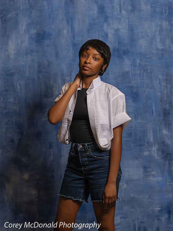 Vivid royal blue abstract hand-painted photography backdrop shown in a studio senior portrait with a young woman in a white shirt and denim shorts, one hand resting at her neck OM-2639
