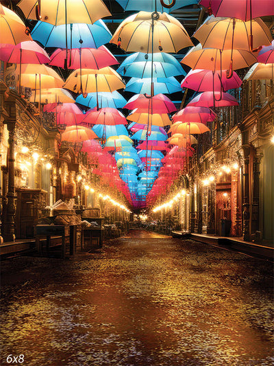 Colorful Umbrella Alley Photography Backdrop - A vibrant alleyway adorned with colorful umbrellas in shades of pink, blue, and yellow, suspended in the air. The scene is illuminated by vintage-style street lamps, creating a whimsical and enchanting atmosphere.