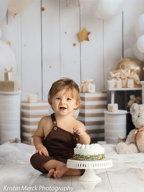 Elegant cake smash photography backdrop featuring a dreamy arrangement of gold, white, and beige balloons, plush teddy bears, stacked presents, and a soft white background with golden stars.
