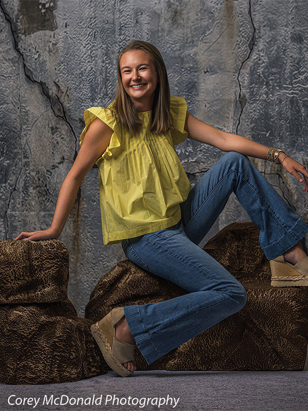Faux rock set photo prop of textured boulders used as studio seating, with a senior girl in a yellow ruffle-sleeve top and blue flared jeans seated on the brown rocks with one foot forward in front of a textured silver industrial wall backdrop.