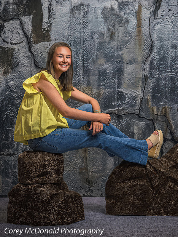 Faux rock set photo prop of textured boulders used as studio seating, with a senior girl in a yellow ruffle-sleeve top and blue flared jeans seated on the brown rocks with legs crossed in front of a textured silver industrial wall backdrop.