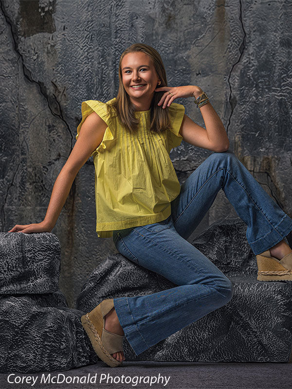 Faux rock set photo prop of textured boulders used as studio seating, with a senior girl in a yellow ruffle-sleeve top and blue flared jeans seated on the grey rocks with a hand near her chin in front of a textured silver industrial wall backdrop.
