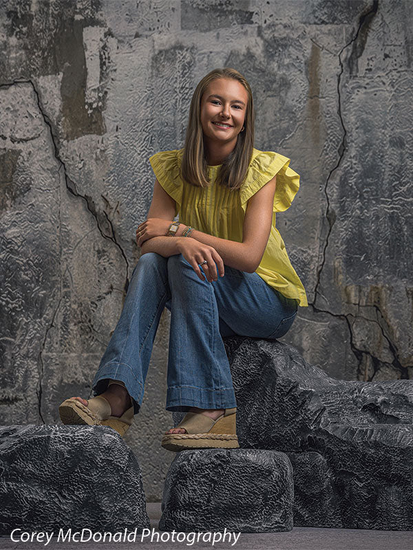 Faux rock set photo prop of textured boulders used as studio seating, with a senior girl in a yellow ruffle-sleeve top and blue flared jeans seated centered on the grey rocks with hands clasped in front of a textured silver industrial wall backdrop.