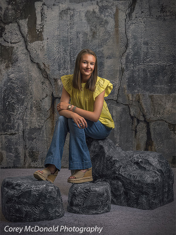 Faux rock set photo prop of textured boulders used as studio seating, with a senior girl in a yellow ruffle-sleeve top and blue flared jeans seated tall on the grey rocks with one foot on a lower rock in front of a textured silver industrial wall backdrop.