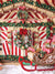 Festive Holiday Gingerbread House Christmas Backdrop - Festive gingerbread house Christmas backdrop featuring a red and white striped facade, green garlands, festive wreaths with ornaments, a decorative sleigh filled with wrapped presents, and fluffy white faux snow on the ground.