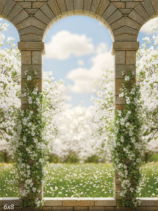 Single stone archway covered in white flowering vines, opening onto a bright spring meadow and blooming orchard beneath a soft blue sky with fluffy clouds.
