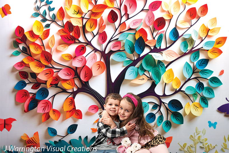 Wide view of rainbow-colored tree filled with vibrant leaves and butterflies, children smiling in front.