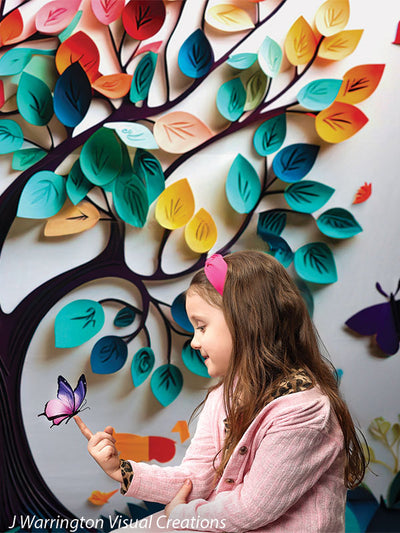 Young girl in pink cardigan admiring a butterfly with vibrant rainbow tree leaves in the background.