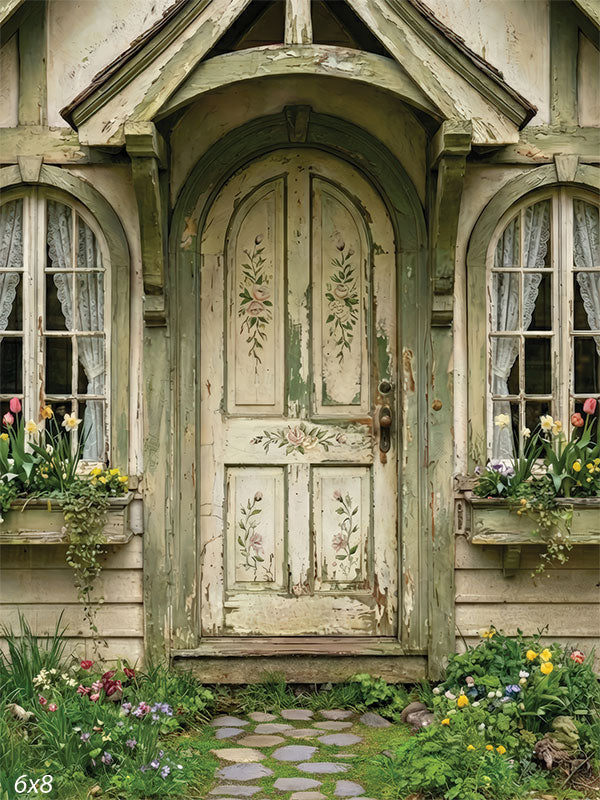 Photography backdrop showing a fairytale cottage façade with an arched wooden door, painted floral details, lace-curtained windows, and garden flowers.