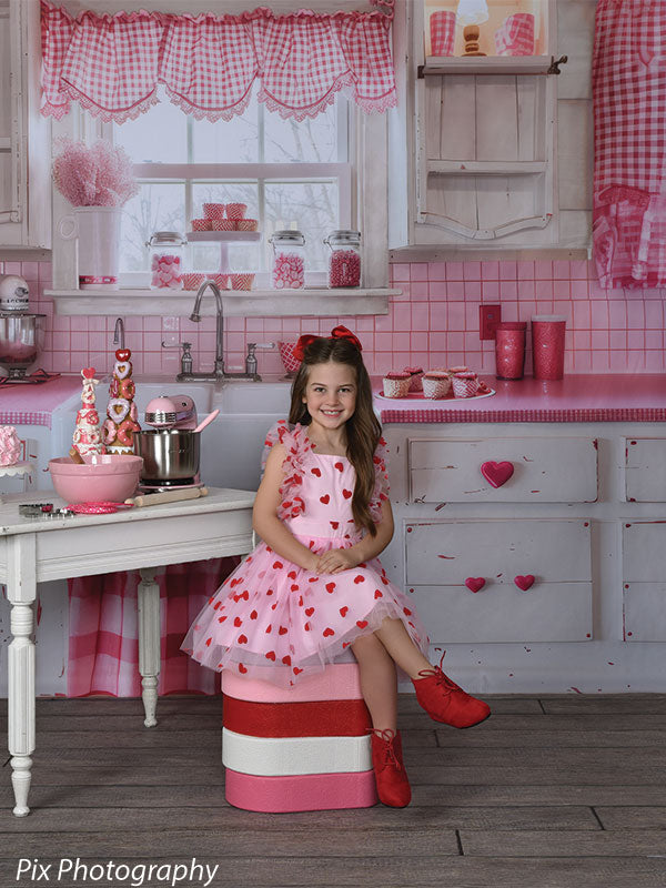 Girl sits on heart-shaped stool prop with pink, red, and white layers in a vintage Valentine kitchen photo scene.