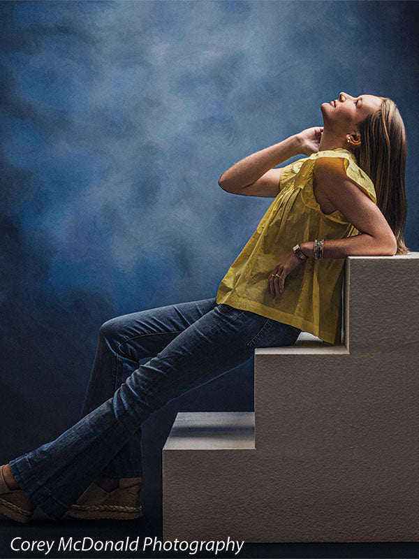 Heavy-duty three-tier foam posing steps used as studio seating, with a senior girl in a yellow ruffle-sleeve top and blue flared jeans leaning back on the foam steps and looking upward against a blue backdrop.