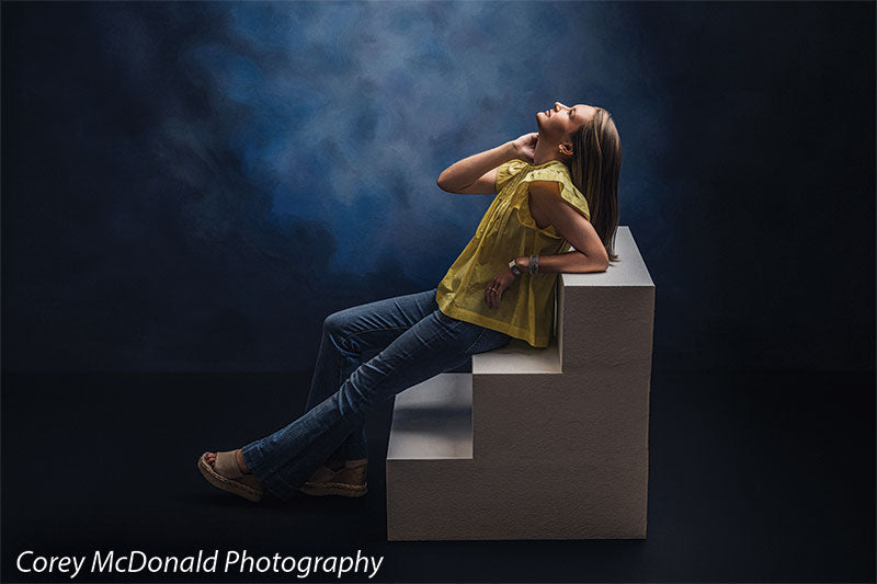 Heavy-duty three-tier foam posing steps used as studio seating, with a senior girl in a yellow ruffle-sleeve top and blue flared jeans reclining against the foam steps looking upward against a blue backdrop.