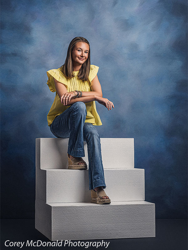 Heavy-duty three-tier foam posing steps used as studio seating, with a senior girl in a yellow ruffle-sleeve top and blue flared jeans seated on the foam steps with one arm resting on her knee against a blue backdrop.