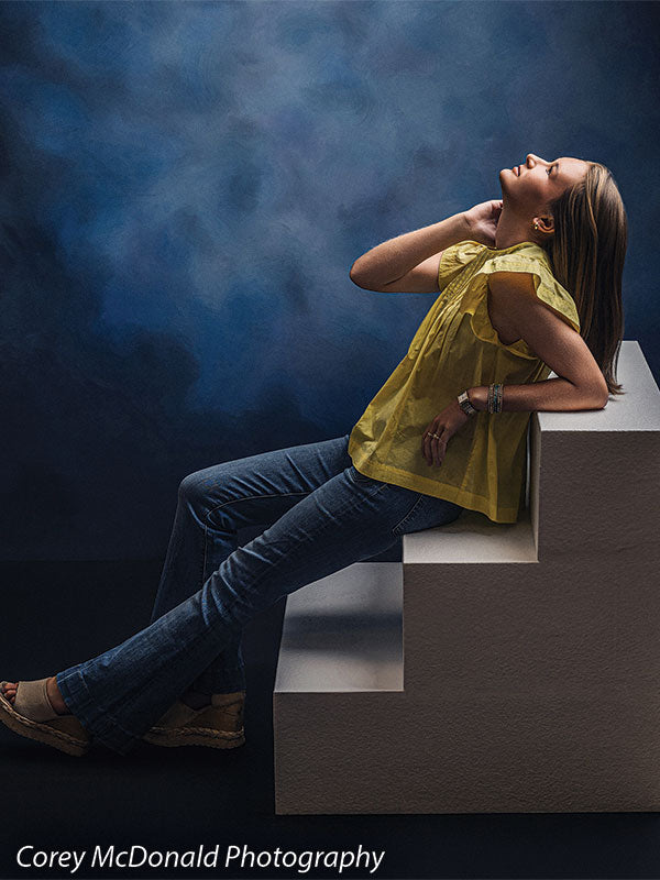 Heavy-duty three-tier foam posing steps used as studio seating, with a senior girl in a yellow ruffle-sleeve top and blue flared jeans seated on the foam steps with a hand near her chin against a blue backdrop.