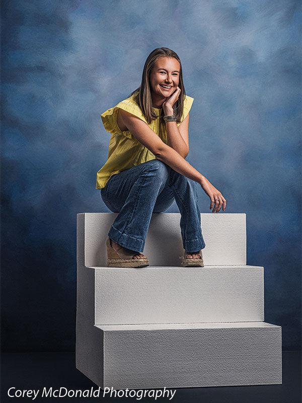 Heavy-duty three-tier foam posing steps used as studio seating, with a senior girl in a yellow ruffle-sleeve top and blue flared jeans seated on the foam steps with a bright smile against a blue backdrop.