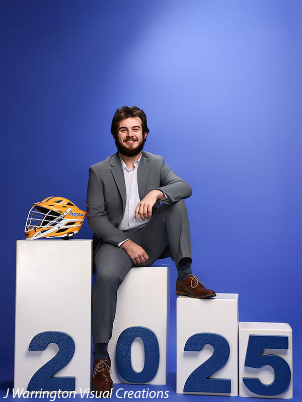 Young man sitting on posing blocks displaying blue magnetic numbers "2025" with a lacrosse helmet as a prop, blue background.