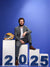 Young man sitting on posing blocks displaying blue magnetic numbers "2025" with a lacrosse helmet as a prop, blue background.
