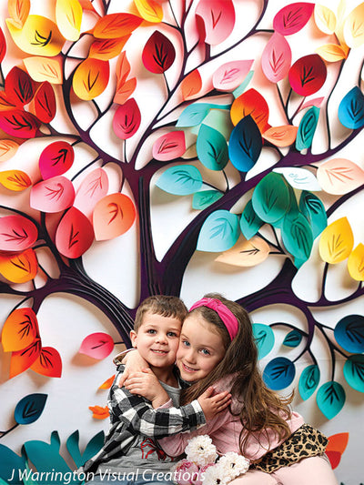 Brother and sister hugging happily in front of a colorful rainbow tree backdrop with vivid paper leaves and butterflies.