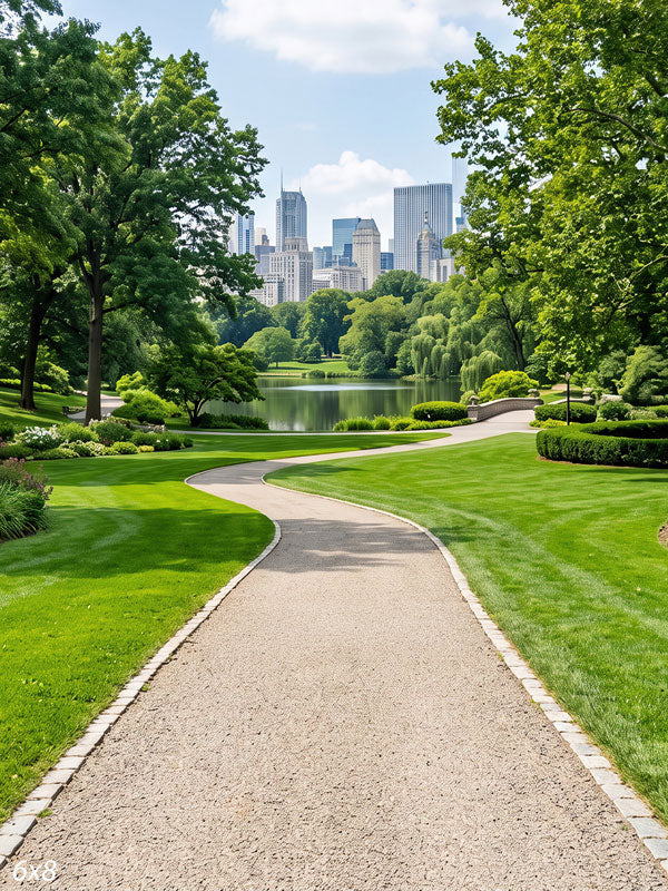 Landscaped city park with a gravel path winding past lawns and flower borders to a lake, with a skyline rising beyond the trees, a scenic photography backdrop.