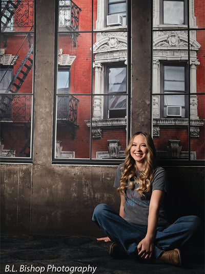 Lifestyle senior photography backdrop with laughing pose against detailed brick building and large windows.