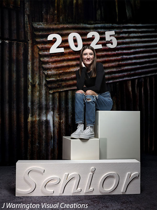 Senior girl seated on posing blocks with white magnetic numbers "2025" on an industrial backdrop. "Senior" displayed on a horizontal block.