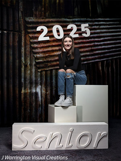 Senior girl seated on posing blocks with white magnetic numbers "2025" on an industrial backdrop. "Senior" displayed on a horizontal block.