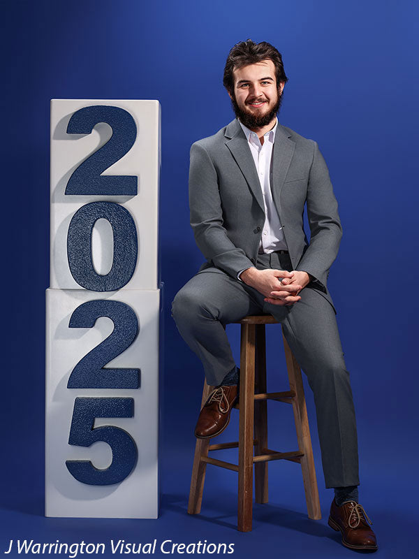 Young man in a gray suit sitting on a stool next to white posing blocks with blue magnetic numbers "2025" on a solid blue background.