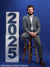 Young man in a gray suit sitting on a stool next to white posing blocks with blue magnetic numbers "2025" on a solid blue background.