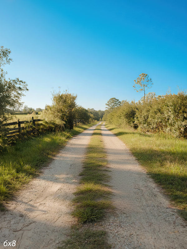Sunny rural dirt lane receding between green grass, trees, and a split-rail fence under a bright blue sky, an outdoor photography backdrop.