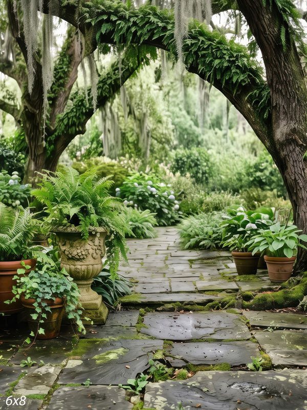 Mossy stone path beneath live oaks draped in Spanish moss with ferns, hostas and carved stone urns, a scenic photography backdrop.