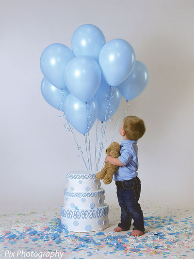 Boy looks up at floating blue balloons in front of a seamless gray backdrop during a styled birthday shoot.