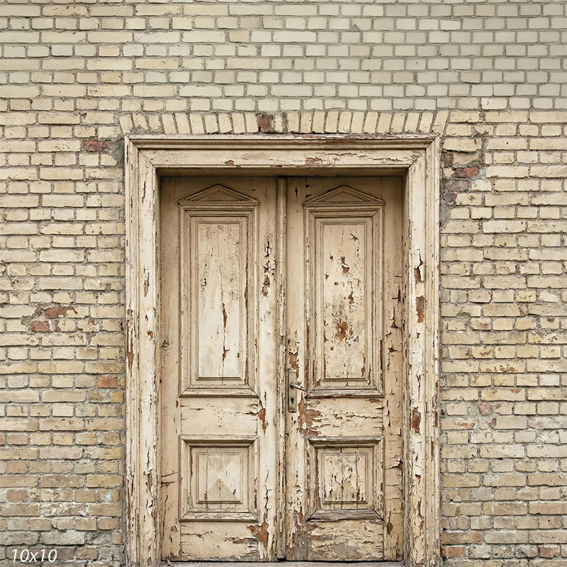 This studio background shows aged double wooden doors with peeling cream paint set into a light brick wall, providing a neutral textured backdrop for portrait photography.