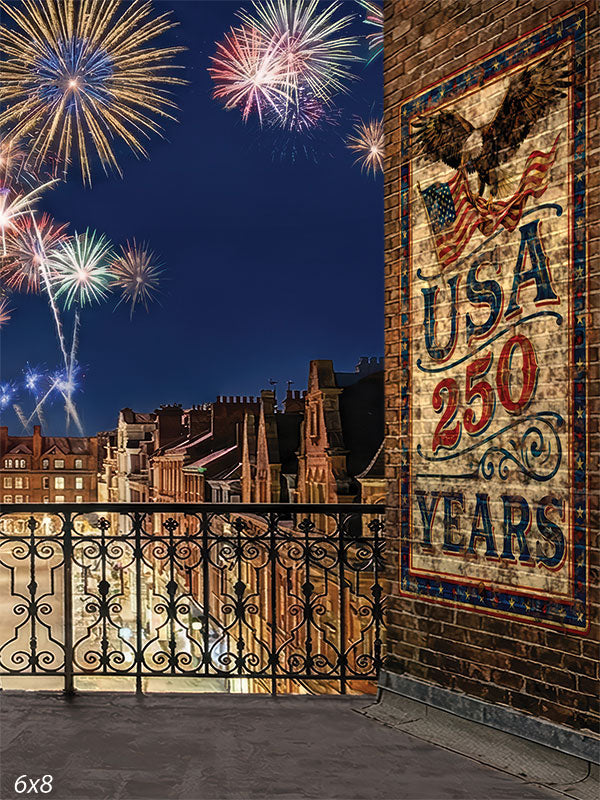 Printed photography backdrop showing a city rooftop with an iron railing, brick wall, and a large USA 250 graphic. Fireworks fill the night sky above the surrounding buildings, creating a patriotic studio background.