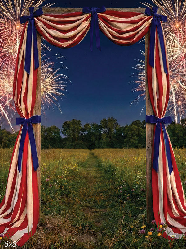 Printed photography backdrop showing a weathered wooden arch draped in red, cream, and blue fabric over a meadow path at night. Fireworks fill the sky behind the arch, with trees and wild grass visible in the background.
